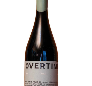 A dark glass wine bottle with a pale green capsule and a white label reading 'OVERTIME' stands on a wooden surface against a wooden background.