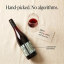 Wine bottle with a glass of red wine and a note on a textured surface, with text 'Hand-picked. No algorithms.'